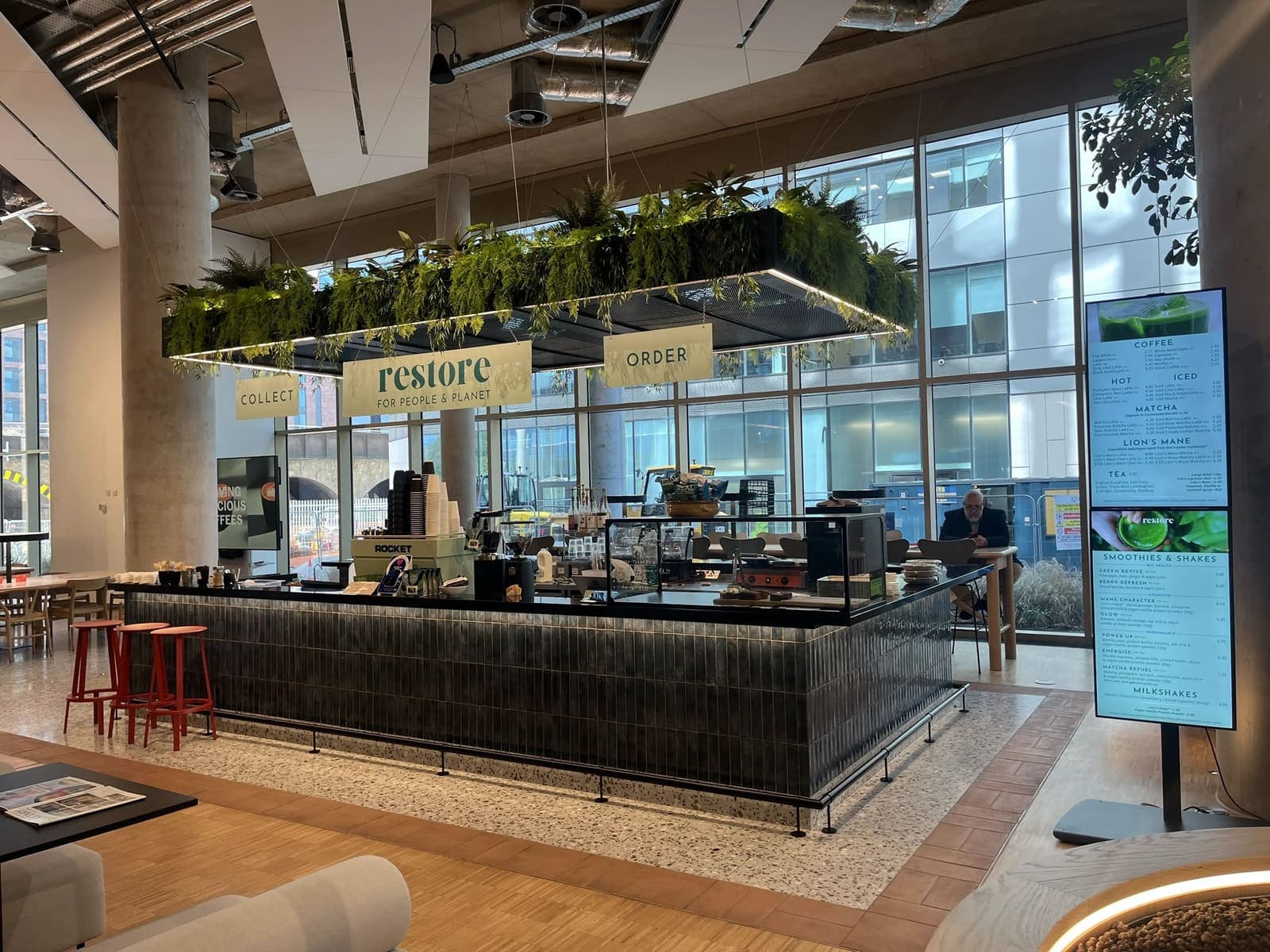 The Restore coffee counter inside the Welcome Building in Central Bristol, with hanging planting above the bar and the menu on a digital display